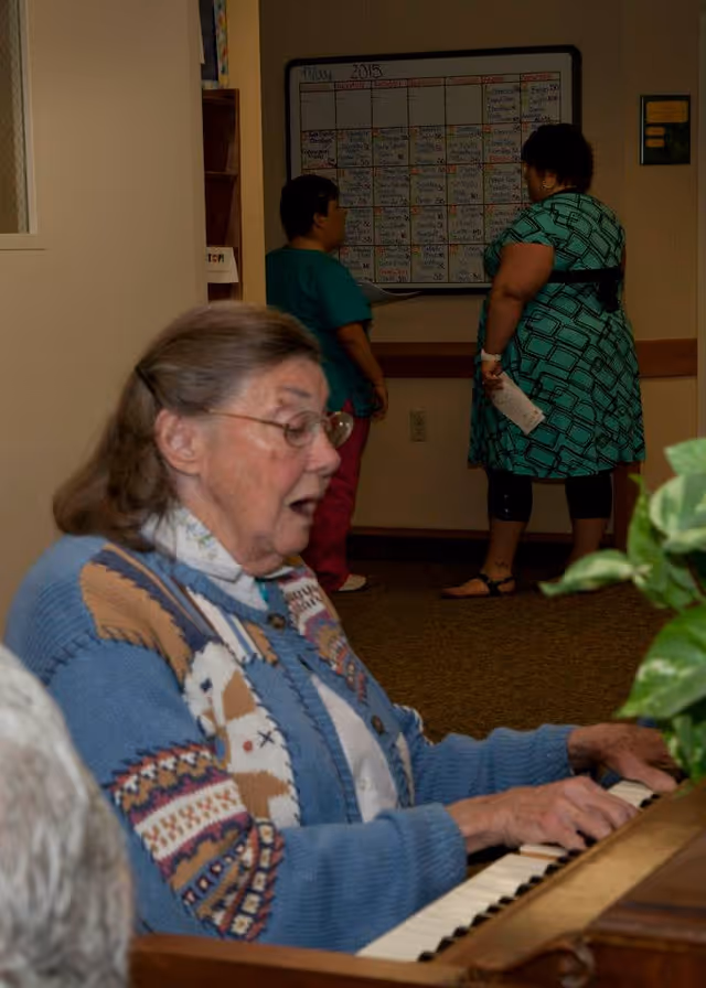 An elderly woman wearing glasses and a patterned blue sweater is playing a piano indoors. In the background, two women stand facing a large whiteboard calendar on the wall, engaged in conversation. The setting appears to be a communal area in a senior living facility.