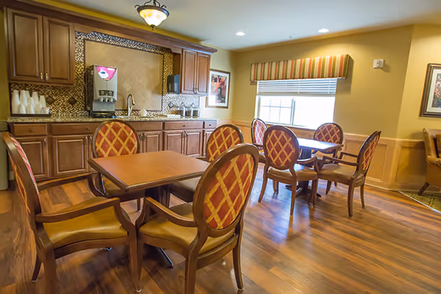 A dining area in a senior living facility with wooden tables and chairs featuring red and yellow patterned upholstery. The room has wooden flooring, a window with a striped valance, and a kitchenette area with cabinets, a coffee machine, and a sink.