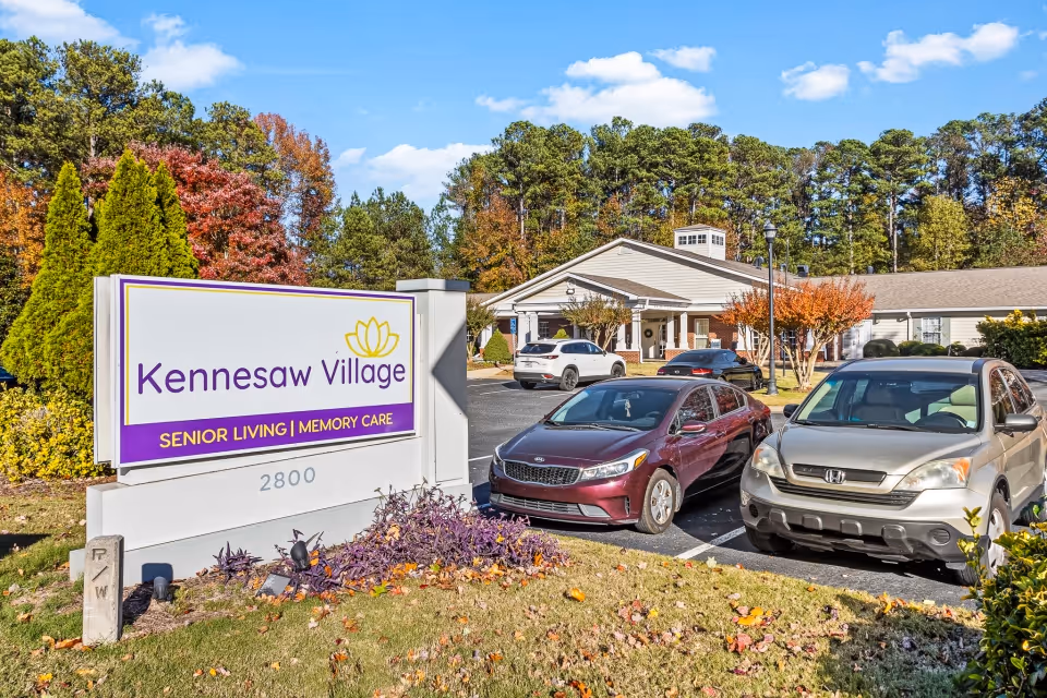 Exterior view of Kennesaw Village Senior Living and Memory Care facility with a large sign in the foreground and several parked cars in front of a single-story building surrounded by trees with autumn foliage.