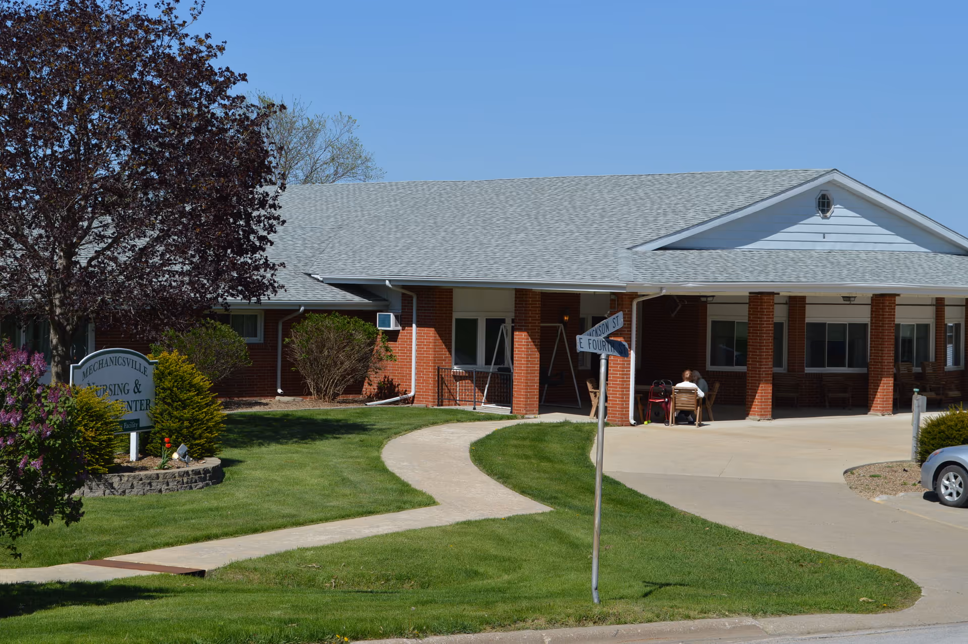Front entrance of the red-brick Mechanicsville Specialty Care nursing center with a curved walkway, landscaped lawn, and a person seated near the covered entry.