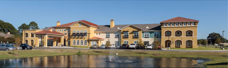 Wide exterior view of The Blake at Lafayette senior living facility showing a large yellow building with multiple windows, balconies, and a red-tiled roof. In front of the building is a pond with a small fountain, a grassy area, and a parking lot with several cars. Trees and a clear blue sky are visible in the background.