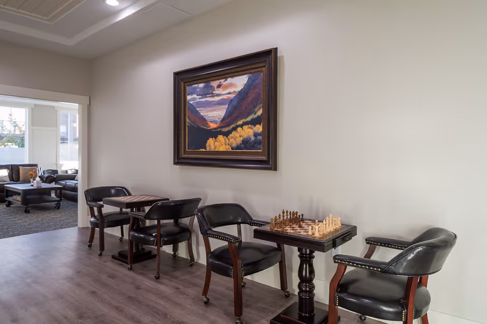 A cozy senior living common area with two small tables and four black leather chairs. One table has a chessboard set up for a game. A framed painting of a mountain landscape with autumn trees hangs on the wall above the tables. In the background, there is a living room area with sofas and a coffee table near large windows letting in natural light.