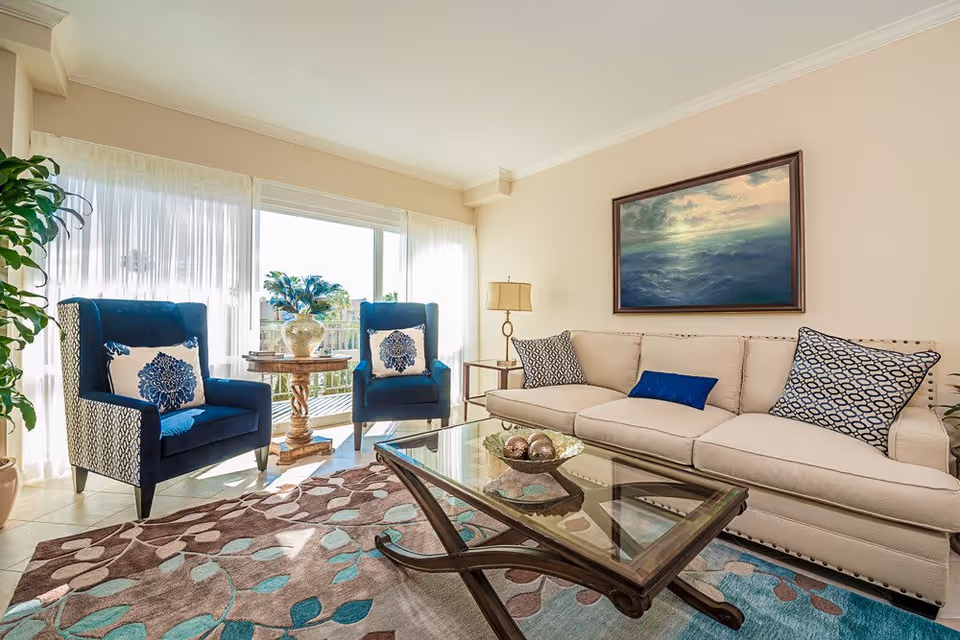 A bright living room with a beige sofa adorned with patterned and solid blue pillows, two blue armchairs with floral cushions, a glass coffee table with decorative items, a wooden side table with a lamp, and a large window with sheer white curtains letting in natural light. A framed painting of a seascape hangs on the wall above the sofa.