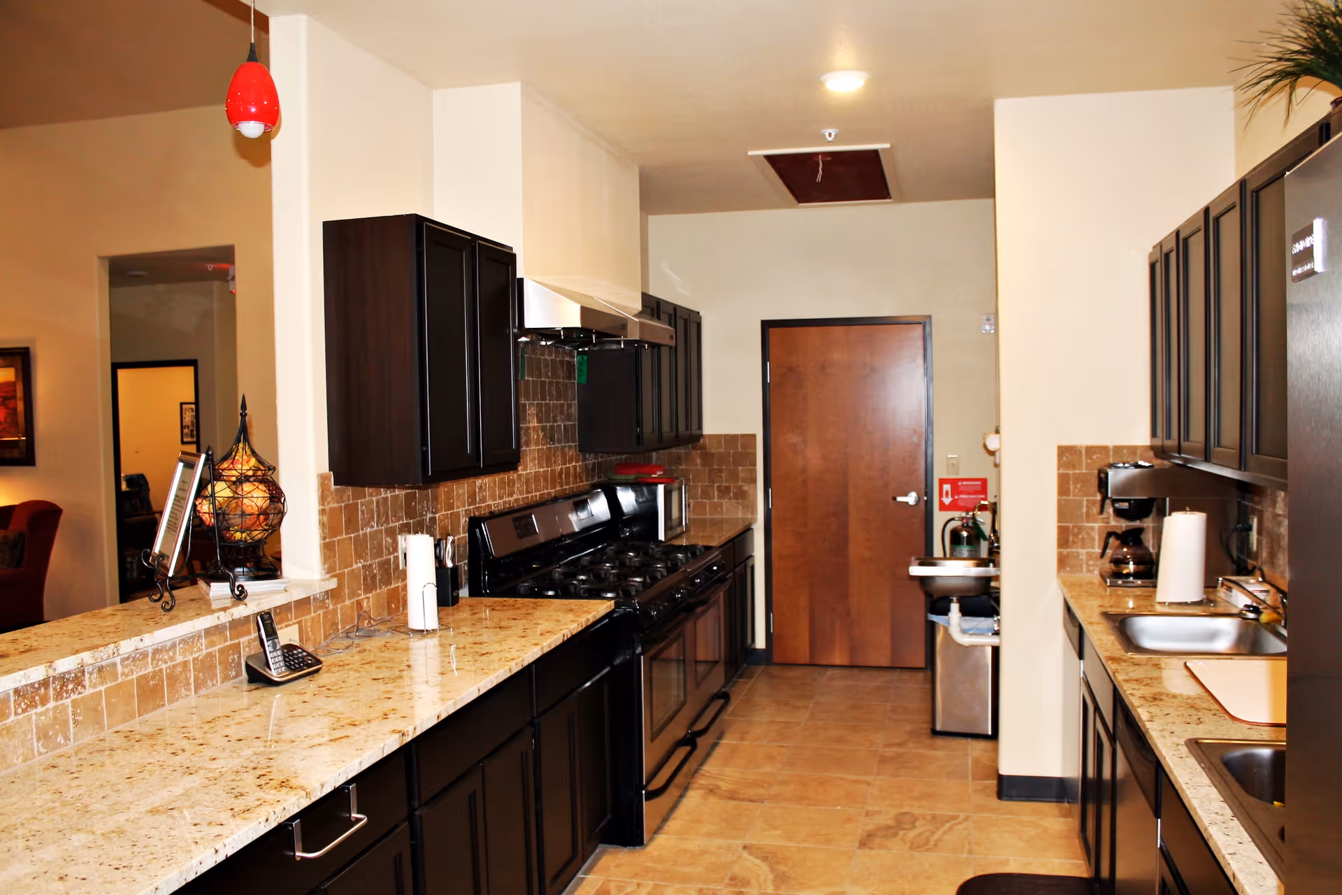 A modern kitchen with dark wood cabinets, granite countertops, a stainless steel stove with a range hood, a refrigerator, a coffee maker, a sink, and a wooden door at the far end. The kitchen has beige tiled floors and walls with a tiled backsplash. A red pendant light hangs from the ceiling on the left side.