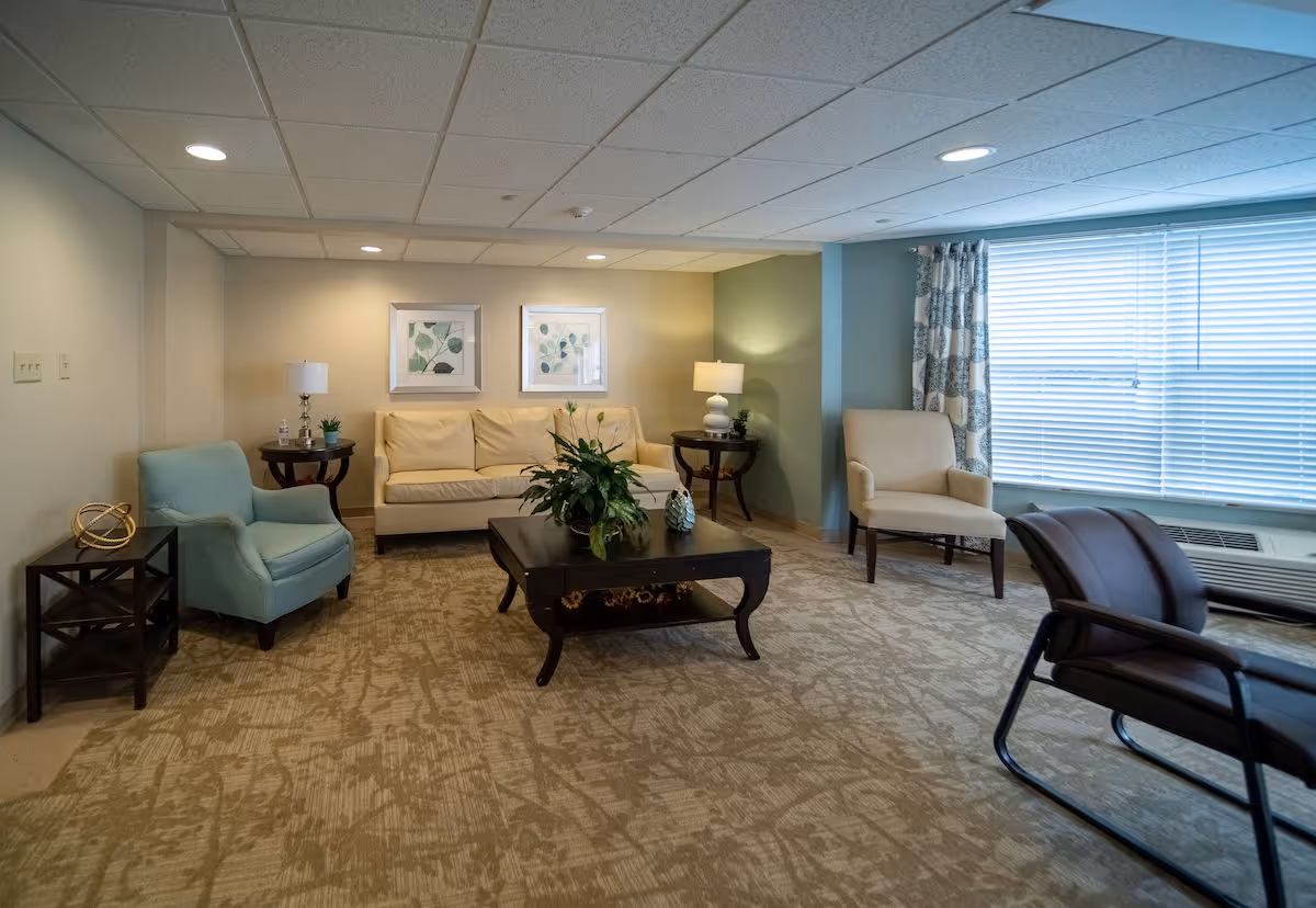 A cozy living room with a beige sofa, two framed botanical prints on the wall, a dark wooden coffee table with a plant centerpiece, two side tables with lamps, a light blue armchair, a beige armchair near a large window with blinds and patterned curtains, and a dark brown chair with armrests.