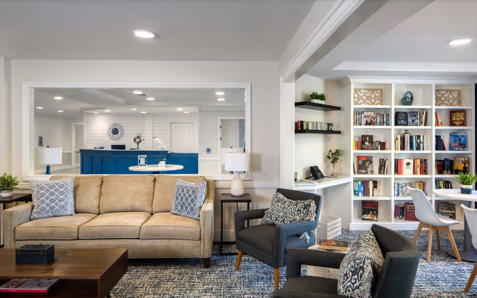 A cozy senior living facility common area with a beige sofa adorned with patterned blue and white pillows, two dark gray armchairs with matching pillows, a wooden coffee table, and a side table with a lamp. In the background, there is a blue reception desk with a clock on the wall and a white built-in bookshelf filled with books and decorative items. The room has a blue patterned carpet and bright ceiling lights.