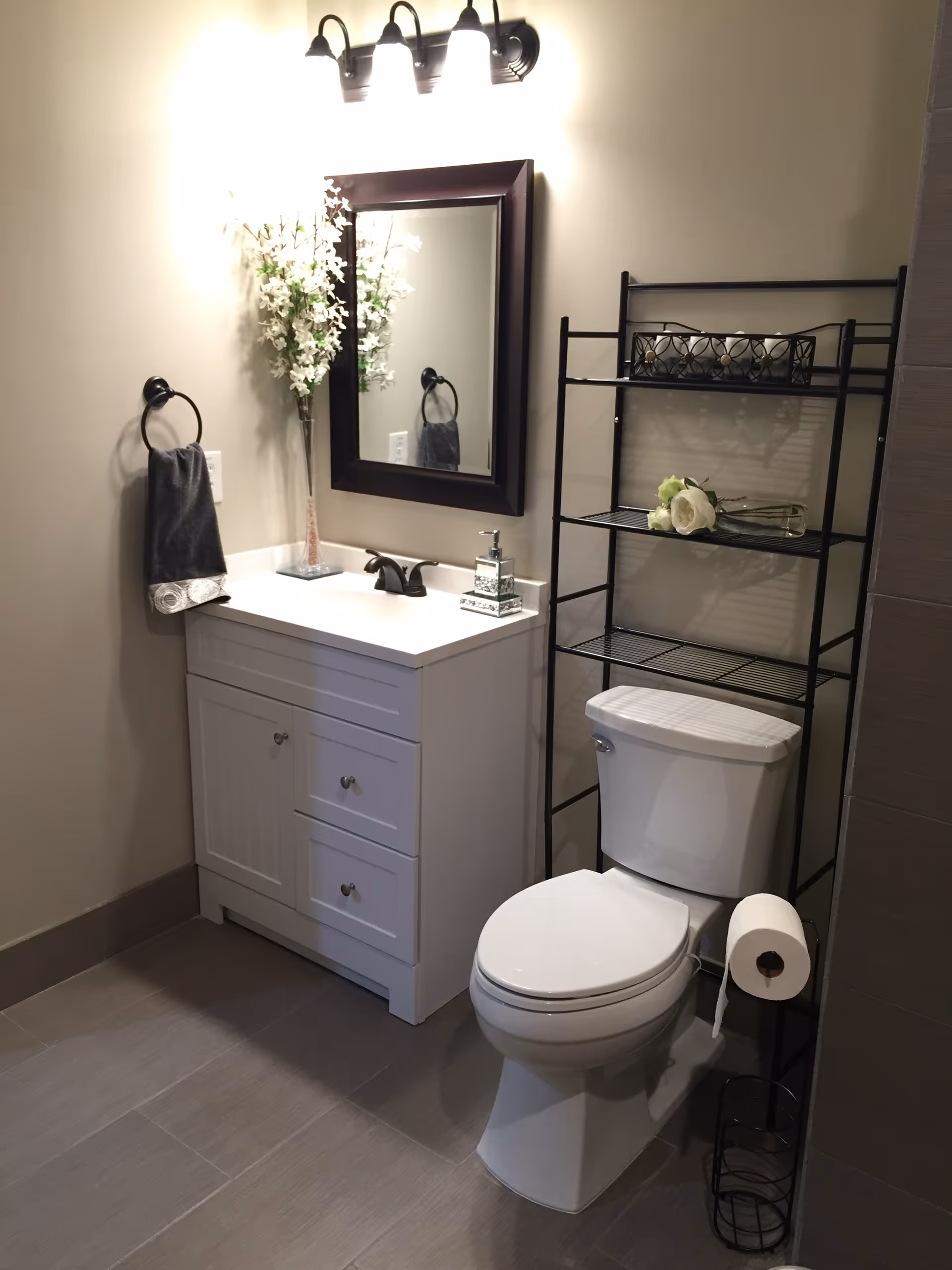Bathroom with a white vanity and sink, mirror and light fixture, toilet, and a black metal shelving unit.