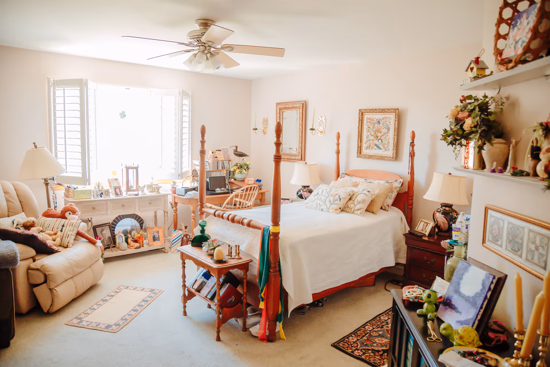 A cozy bedroom with a wooden four-poster bed covered with white bedding and multiple pillows. The room has a beige carpet and walls decorated with framed artwork. There is a beige recliner chair with stuffed animals on it near a window with white shutters. A wooden desk with a chair and various items is positioned near the window. The room also features a ceiling fan with lights, a small wooden table at the foot of the bed, and shelves with decorative items and plants.