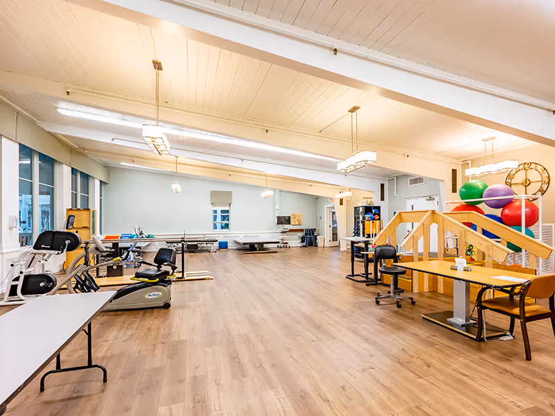 A spacious rehabilitation or therapy room with exercise equipment, therapy tables, colorful exercise balls, and wooden stairs. The room has large windows on one side, light wood flooring, and bright overhead lighting.