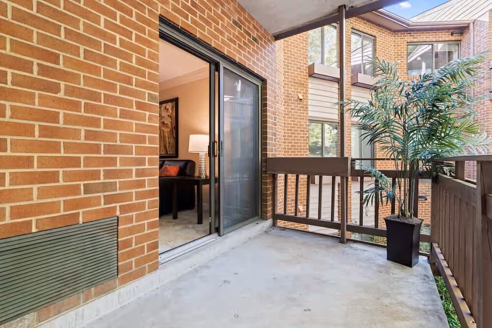Concrete covered balcony with wooden railing and a potted plant, opening through a sliding glass door to a living room against brick exterior walls.