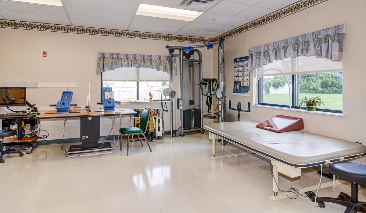 A therapy or rehabilitation room with exercise equipment, a padded treatment table, chairs, and a desk with computer monitors. The room has two windows with floral valances and a view of green grass outside.