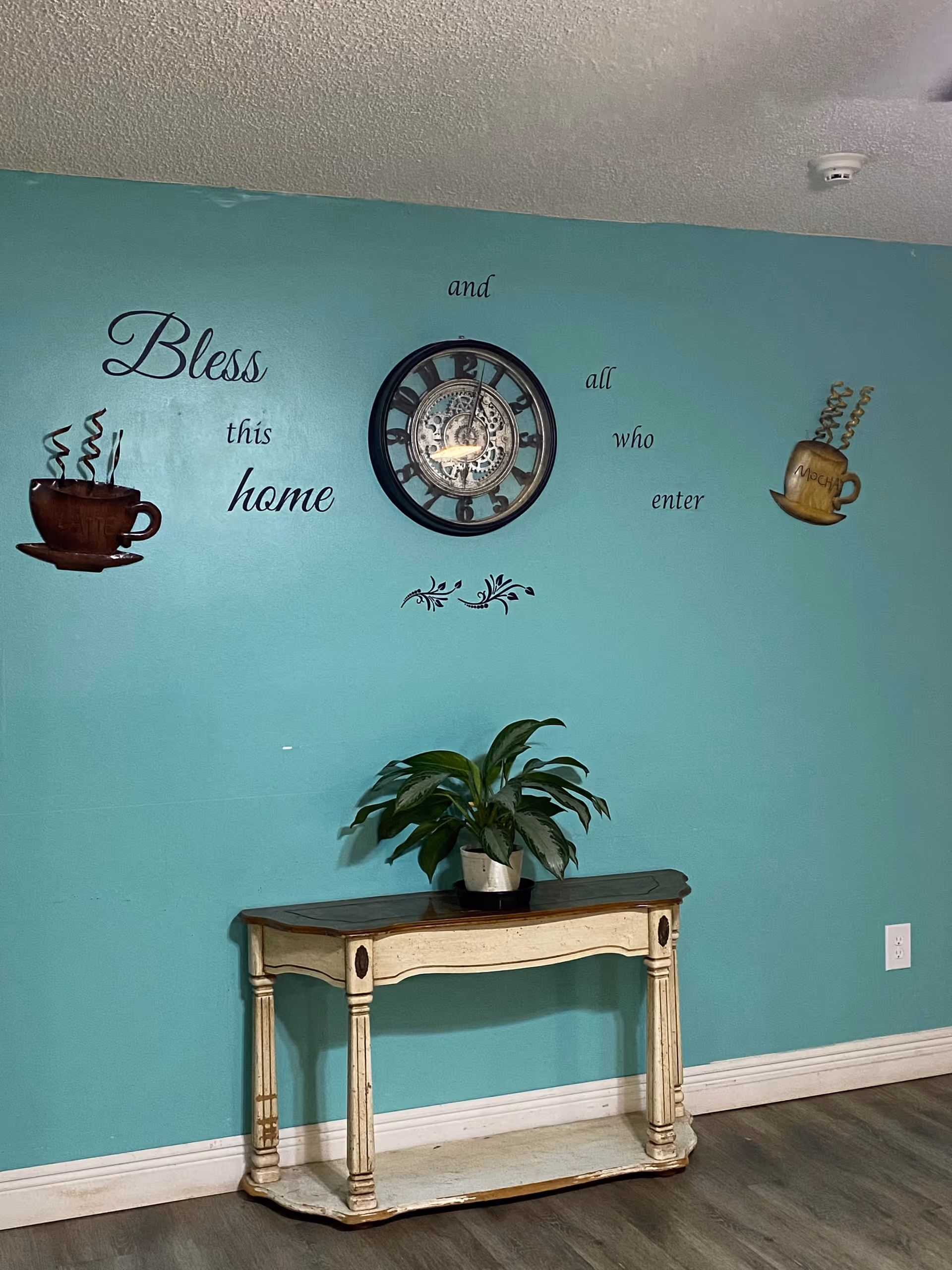 A teal-colored wall with a decorative clock in the center and wall art featuring two steaming coffee cups on either side. The wall art includes the phrase 'Bless this home and all who enter.' Below the clock is a small wooden console table with a potted green plant on top. The floor is wood with white baseboards.