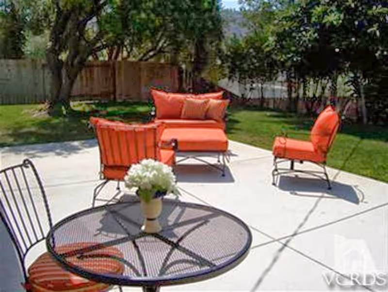 Outdoor patio area with metal table and chairs featuring orange cushions, a vase with white flowers on the table, and additional orange cushioned seating including a loveseat and chairs on a concrete patio surrounded by grass and trees.