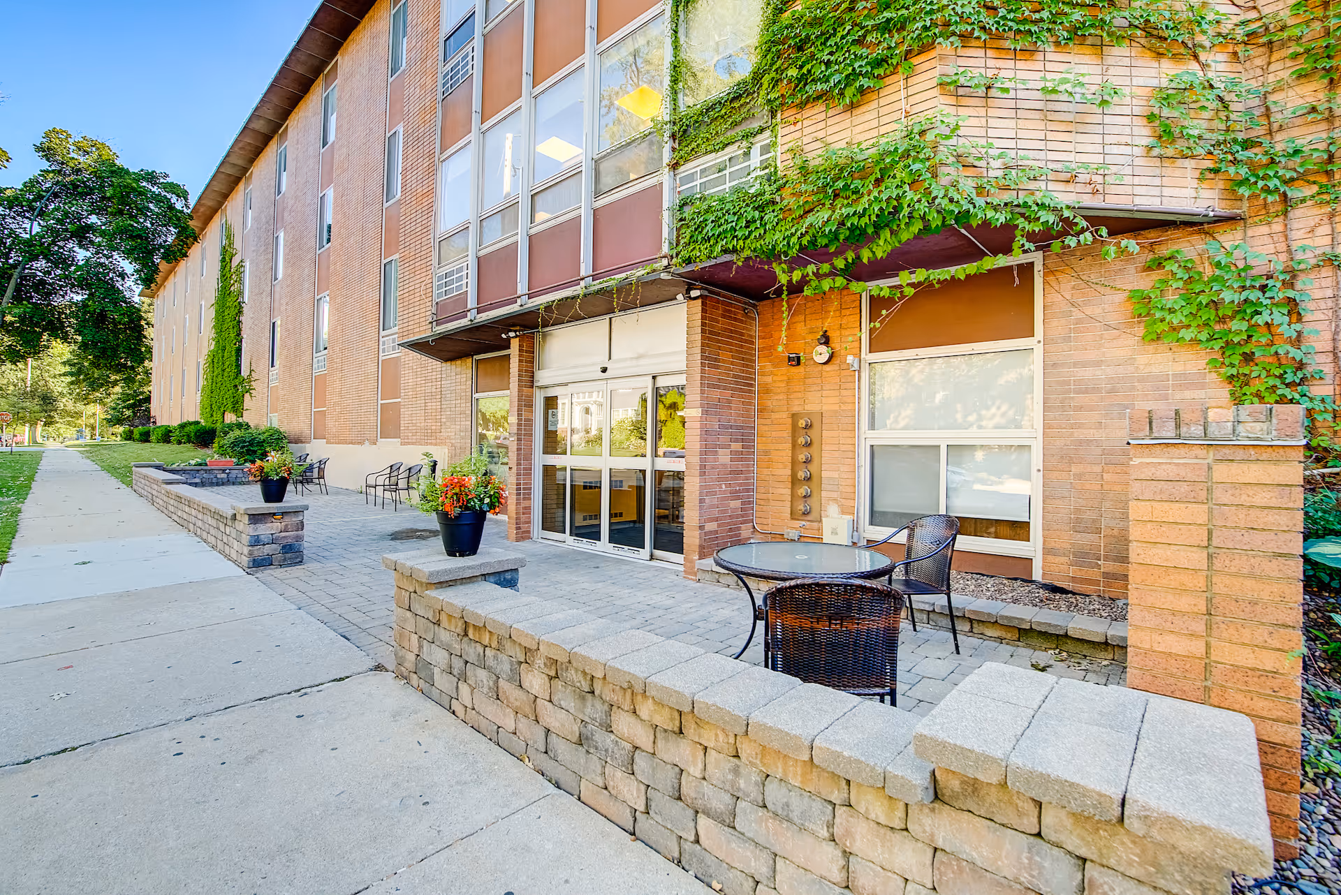 Outdoor patio area of a brick building with climbing ivy on the walls. The patio has a stone wall border, a round glass table with two wicker chairs, and potted plants with flowers. A sidewalk runs alongside the building with additional seating further down.
