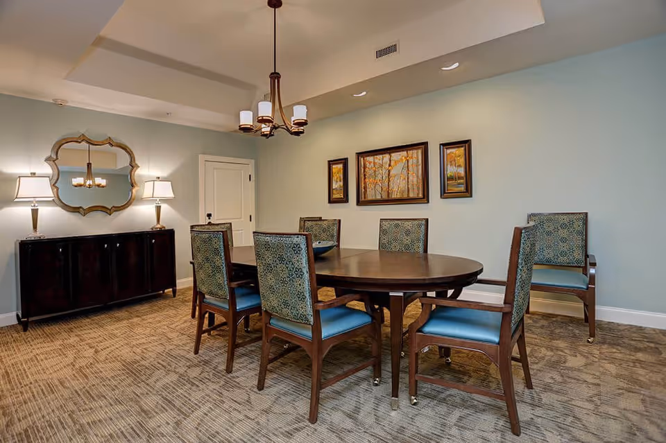 A dining room with a round wooden table surrounded by six chairs with blue patterned upholstery. The room has light blue walls, a beige carpet, and a dark wooden sideboard with two lamps and a decorative mirror above it. Three framed paintings depicting autumn trees hang on the wall behind the table. A chandelier with five lights hangs from the ceiling.
