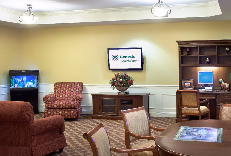 A cozy living room area with patterned armchairs, a wooden cabinet with a floral arrangement, a wall-mounted TV displaying the Genesis HealthCare logo, a fish tank in the corner, and a wooden desk with a computer and chair. The room has beige walls, carpeted floor, and soft ceiling lighting.