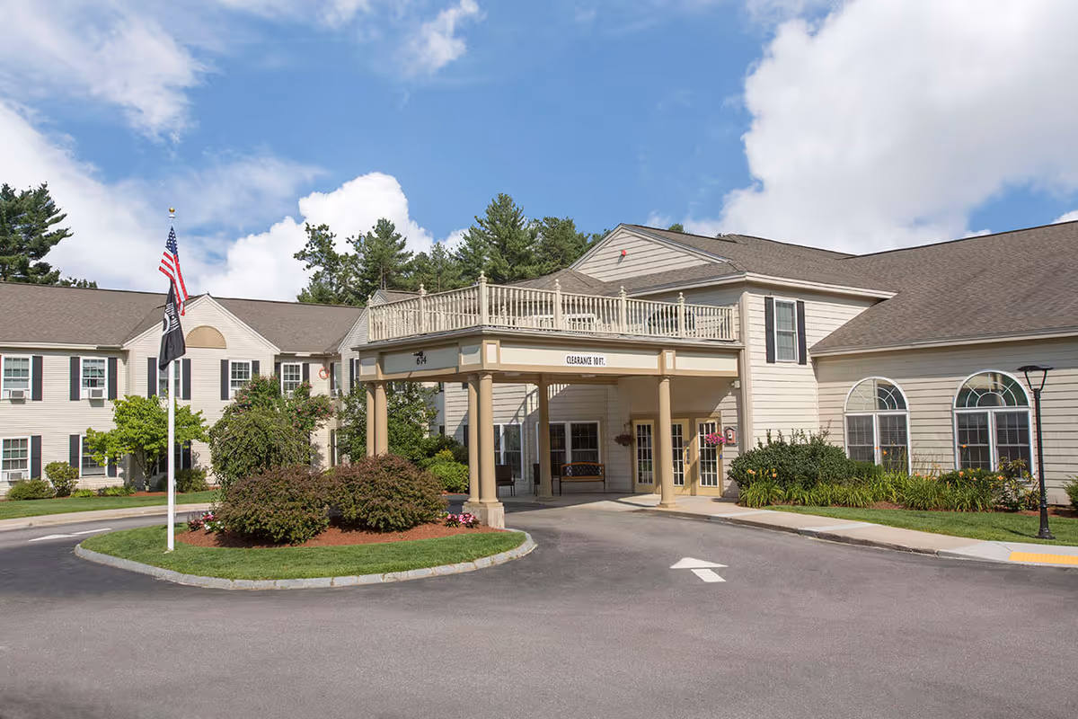 Exterior view of Benchmark Senior Living at Nashua Crossings showing the main entrance with a covered drop-off area supported by columns, landscaped bushes and flowers, American and POW flags on a flagpole, and a clear blue sky with some clouds.