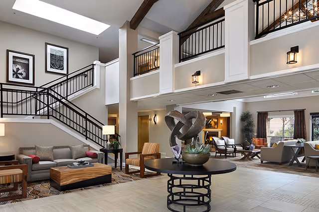 Spacious and well-lit senior living facility common area with a modern staircase featuring black railings, comfortable seating including sofas and armchairs, a round table with a decorative sculpture and plants, and large windows allowing natural light to fill the room.