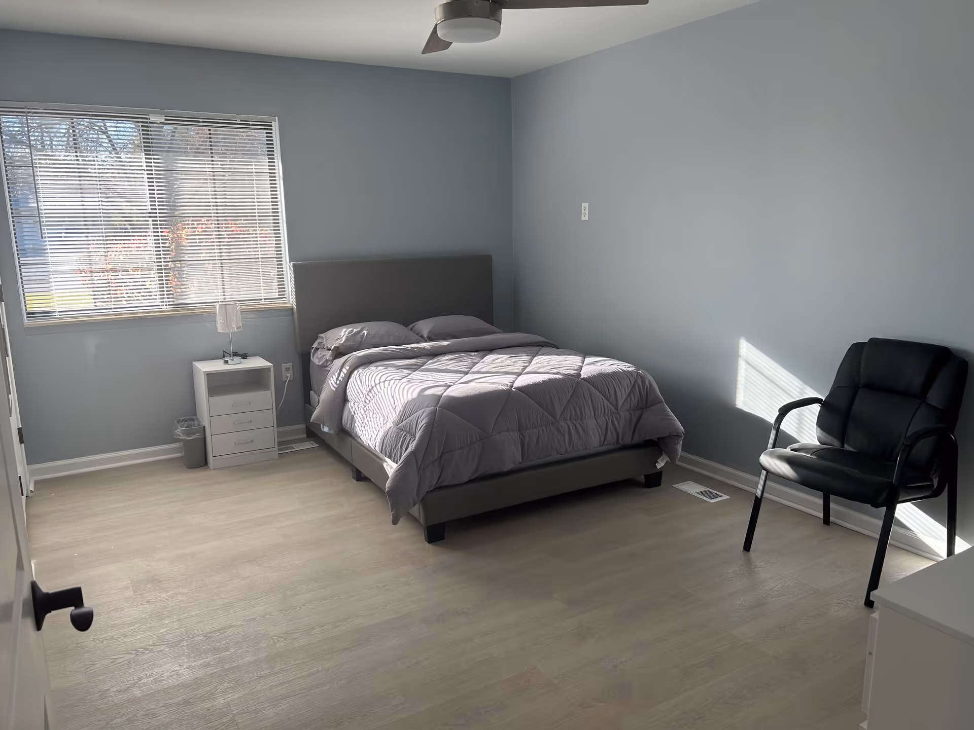 Minimal, sunlit bedroom with a gray bed, nightstand by a window with blinds, and a black chair on light wood flooring.