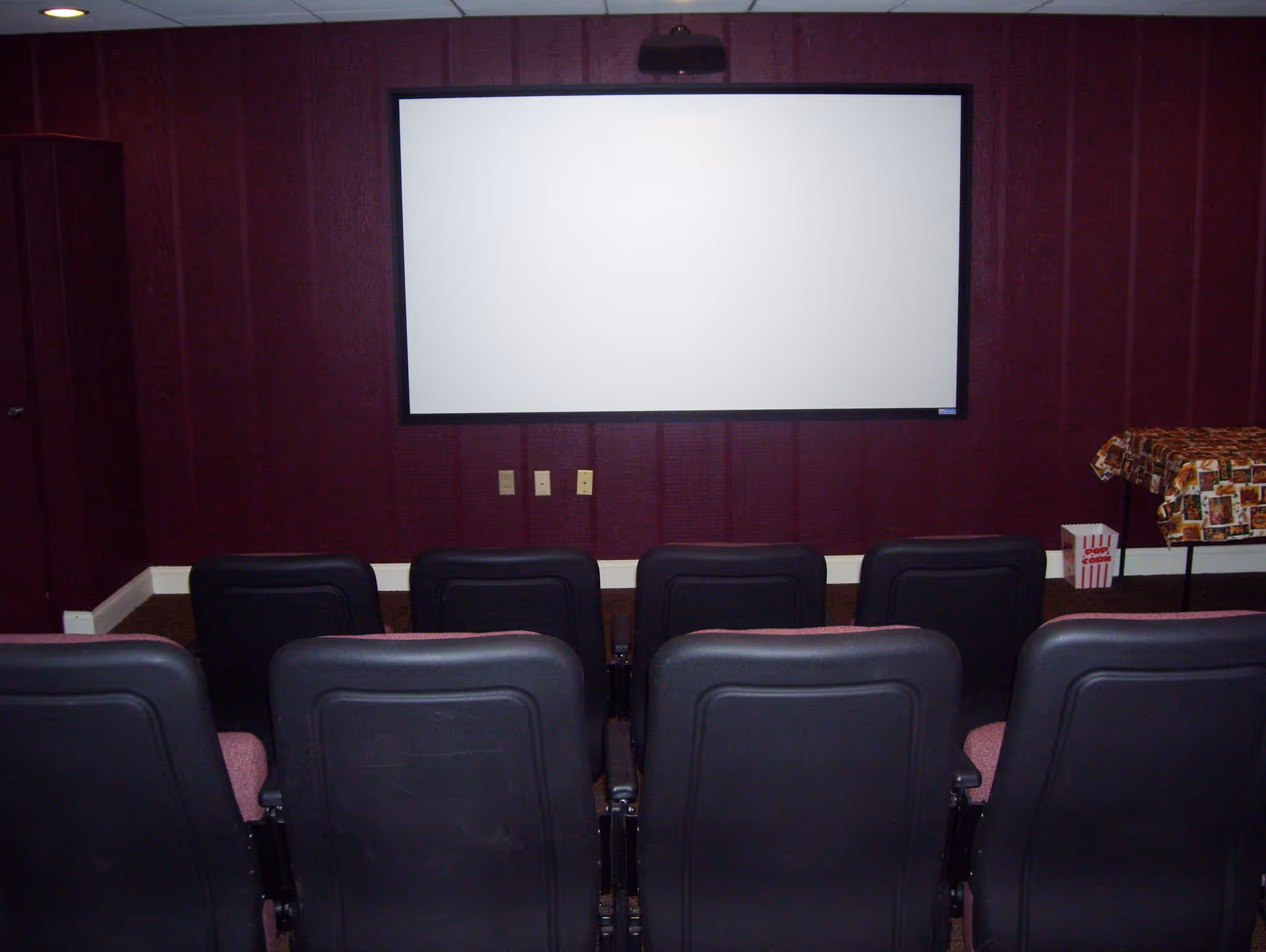 Small movie theater room with a white projection screen on a dark red paneled wall. There are two rows of black and maroon theater seats facing the screen. To the right, there is a table covered with a patterned cloth and a popcorn box on the floor.