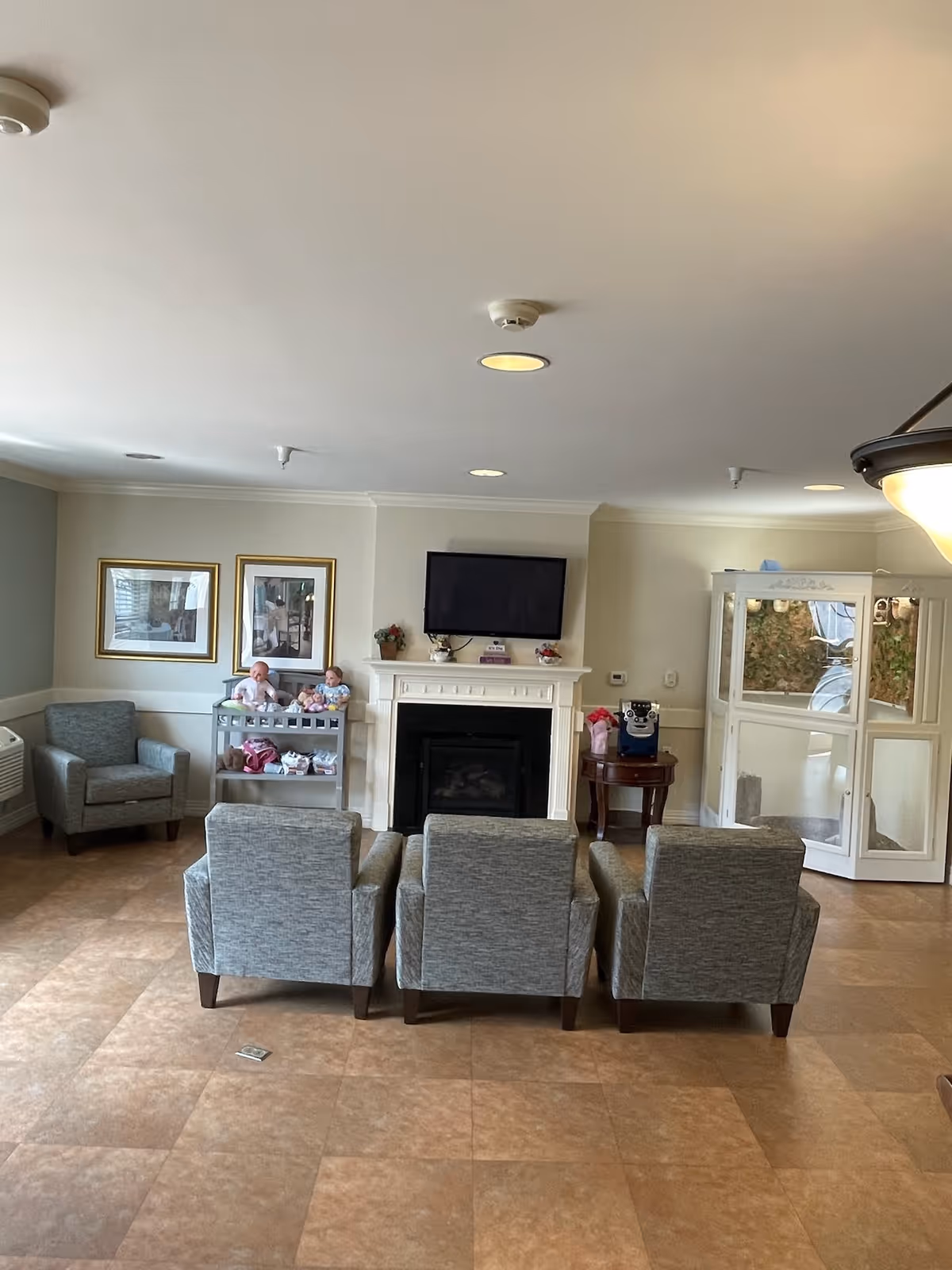 A cozy living room area with three gray armchairs facing a white fireplace with a mounted flat-screen TV above it. To the left, there is a gray armchair next to a small shelf holding dolls and other items. On the right side, there is a small round table with a coffee machine and a white enclosed phone booth. The room has beige tiled flooring and light-colored walls with two framed pictures hanging above the shelf.