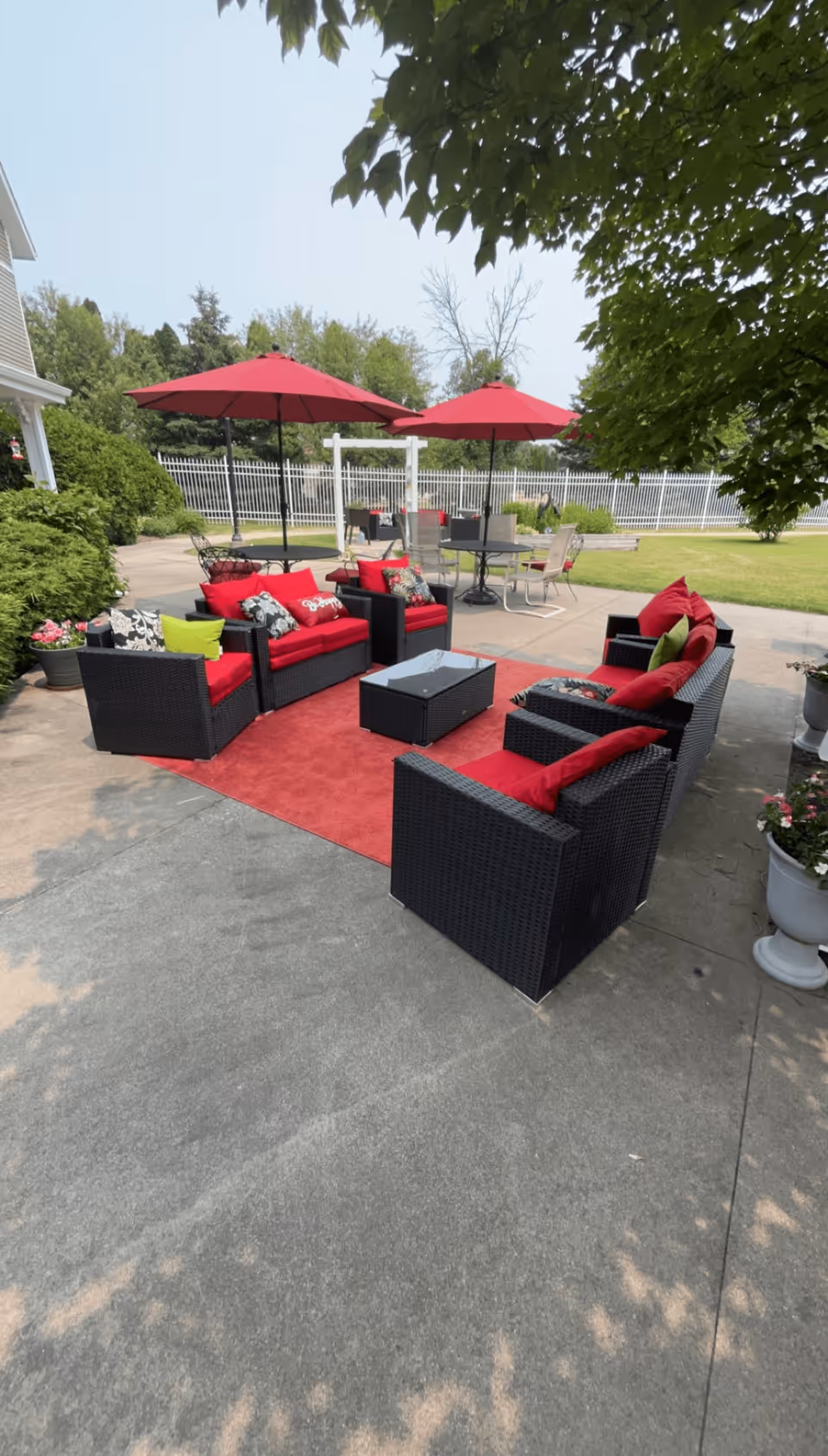 Outdoor patio area with black wicker furniture featuring red cushions and pillows arranged around a rectangular glass-top coffee table on a red outdoor rug. Two red umbrellas provide shade over additional tables and chairs in the background. The area is surrounded by greenery, a white fence, and a clear sky.