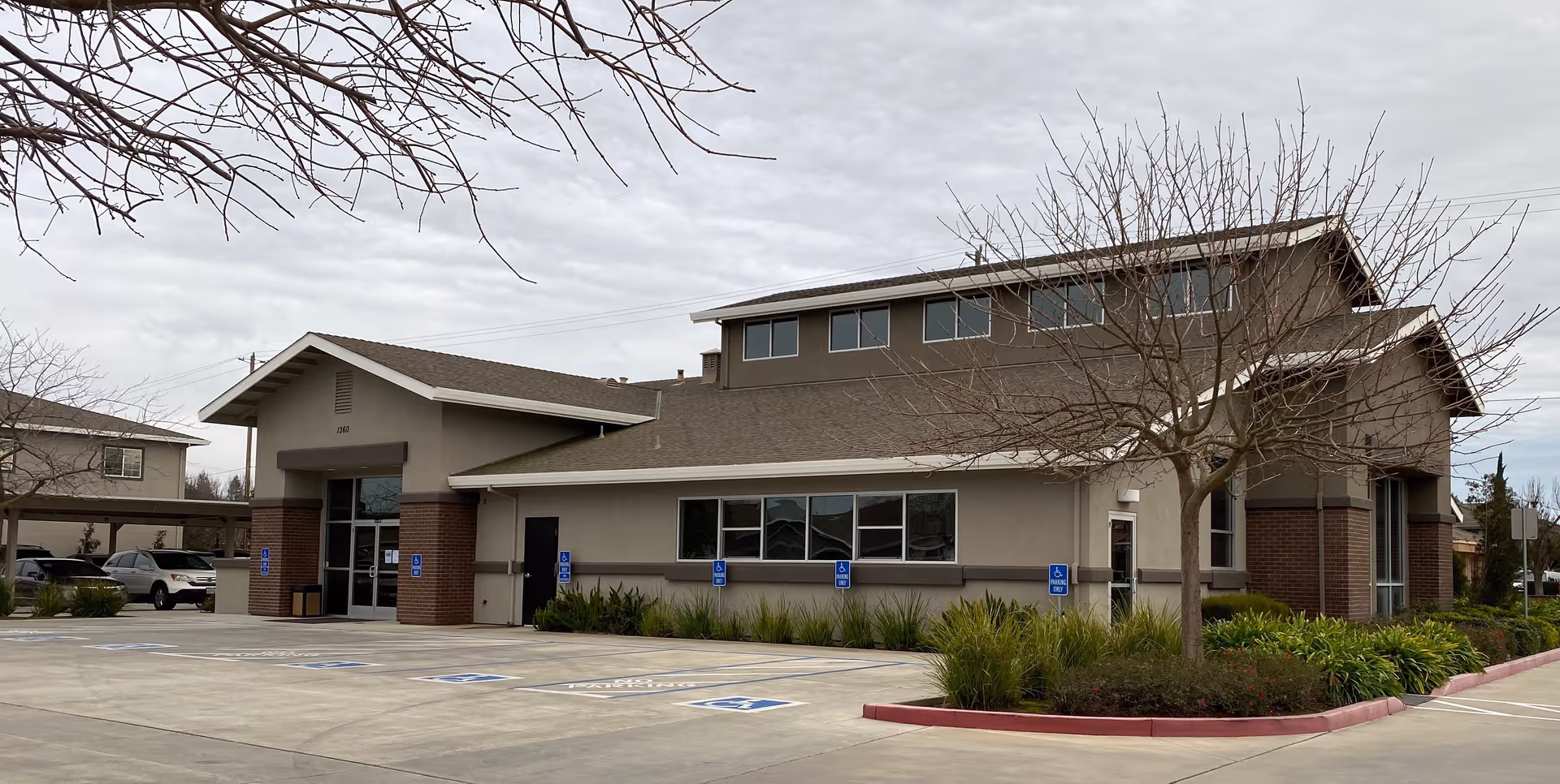 Front exterior of a beige, brick-accented senior living facility with accessible parking spaces and leafless trees in the foreground.
