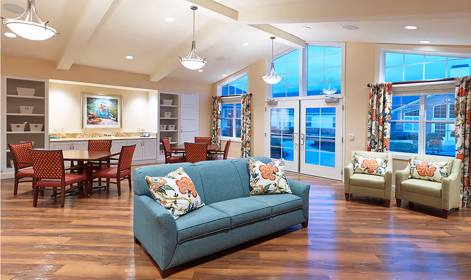 A bright and spacious common area with a teal sofa adorned with floral pillows in the foreground. Behind the sofa, there are two beige armchairs with matching floral pillows near large windows with floral curtains. To the left, there are two wooden tables surrounded by red cushioned chairs. The room features wooden flooring, high ceilings with exposed beams, and pendant lights. A painting and built-in shelves with baskets are visible on the far wall.