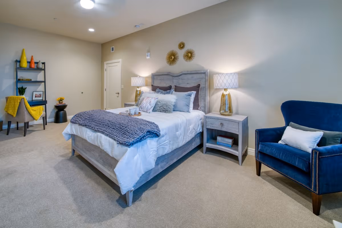 A cozy bedroom with a wooden bed frame, white bedding, and multiple pillows including a fluffy gray one. There are two bedside tables with matching lamps on either side of the bed. A blue upholstered armchair with pillows is placed to the right. On the left side of the room, there is a black shelving unit with decorative items and a beige chair draped with a yellow blanket. The walls are light-colored and decorated with three golden wall ornaments above the bed.