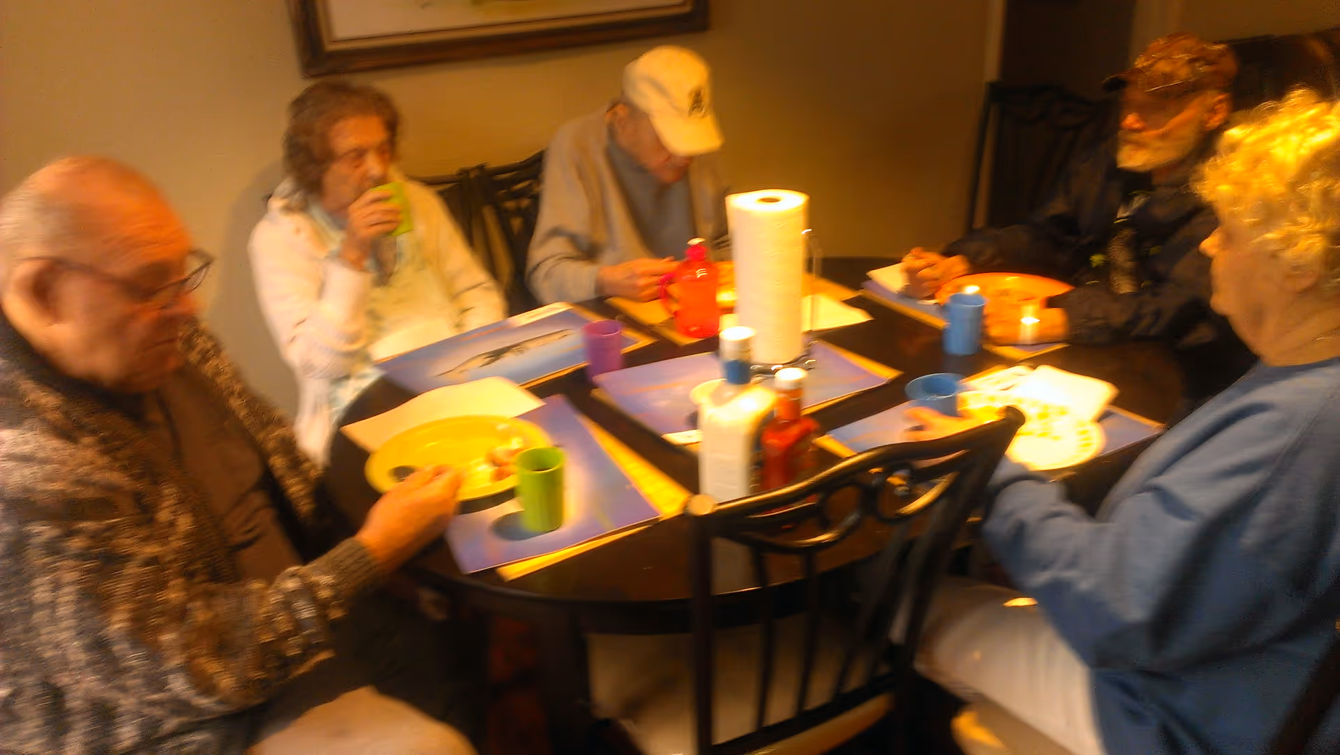 Five elderly residents sit around a round dining table with plates, cups, and condiments.