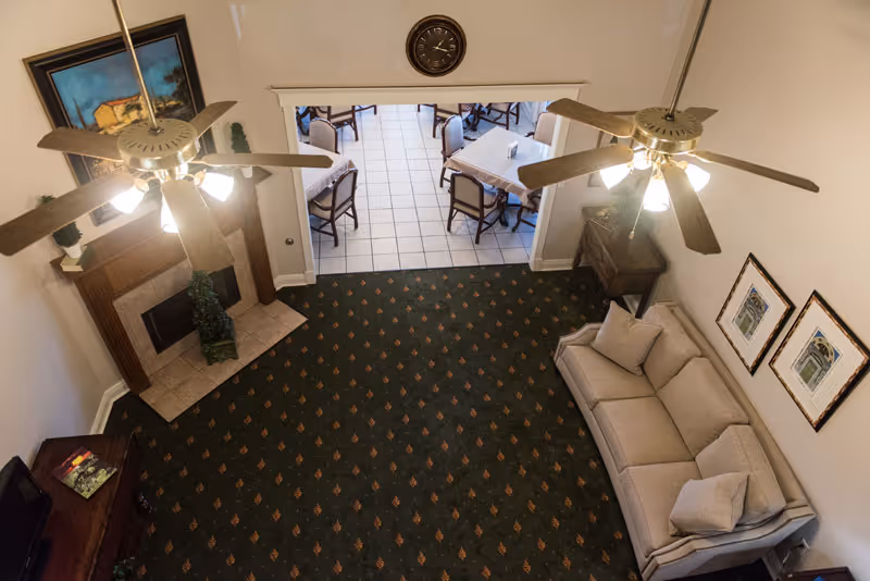 View from above of a living room area with a beige couch, two ceiling fans with lights, a fireplace with a painting above it, and a small table with chairs visible through an open doorway leading to a tiled dining area.
