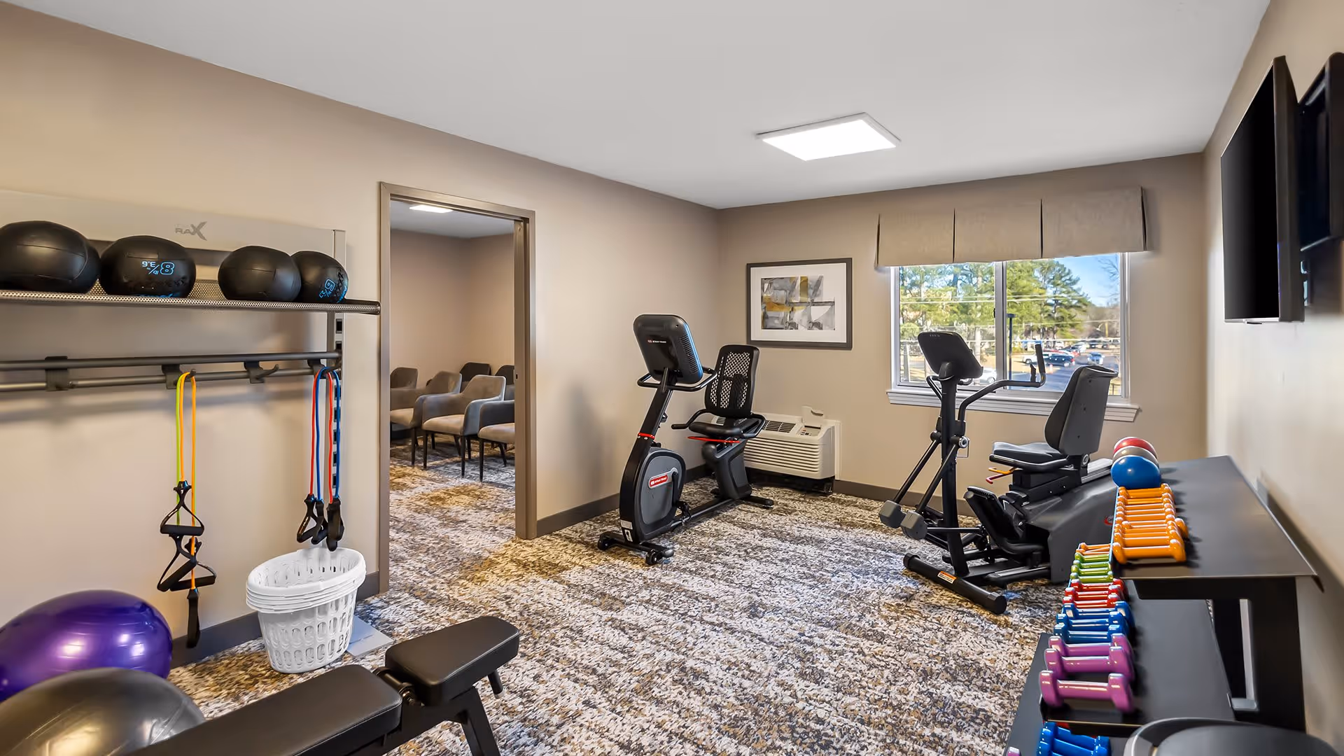A small fitness room with exercise equipment including stationary bikes, resistance bands, medicine balls, dumbbells, and a bench. The room has carpeted flooring, beige walls, a window with a view outside, and a wall-mounted TV. There is an adjacent room visible through an open doorway with several chairs arranged in rows.