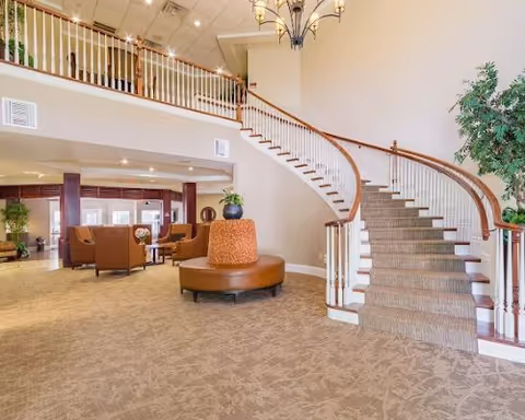 Spacious interior of a senior living facility featuring a curved staircase with carpeted steps and wooden handrails, a round cushioned bench with a plant on top in the center, and a seating area with brown chairs and tables in the background. The space has high ceilings with recessed lighting and a chandelier, beige walls, and carpeted floors.