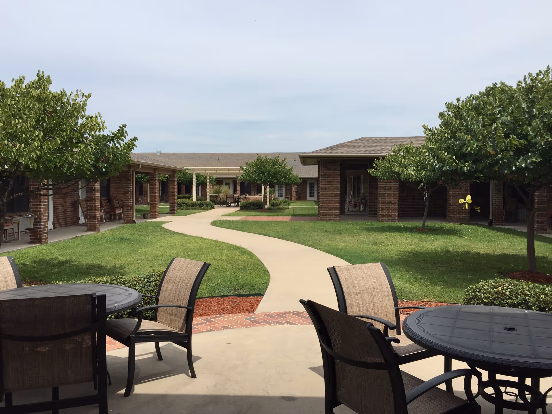 Outdoor courtyard area at Bella Valley Estates featuring a winding concrete pathway surrounded by green grass, small trees, and brick buildings. There are patio tables and chairs in the foreground and rocking chairs along the covered walkways.