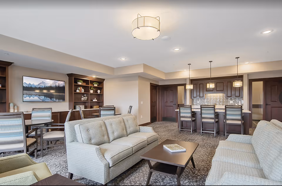Bright common living area with sofas, a coffee table, dining chairs, built-in shelving, and a kitchen-style bar with pendant lights.