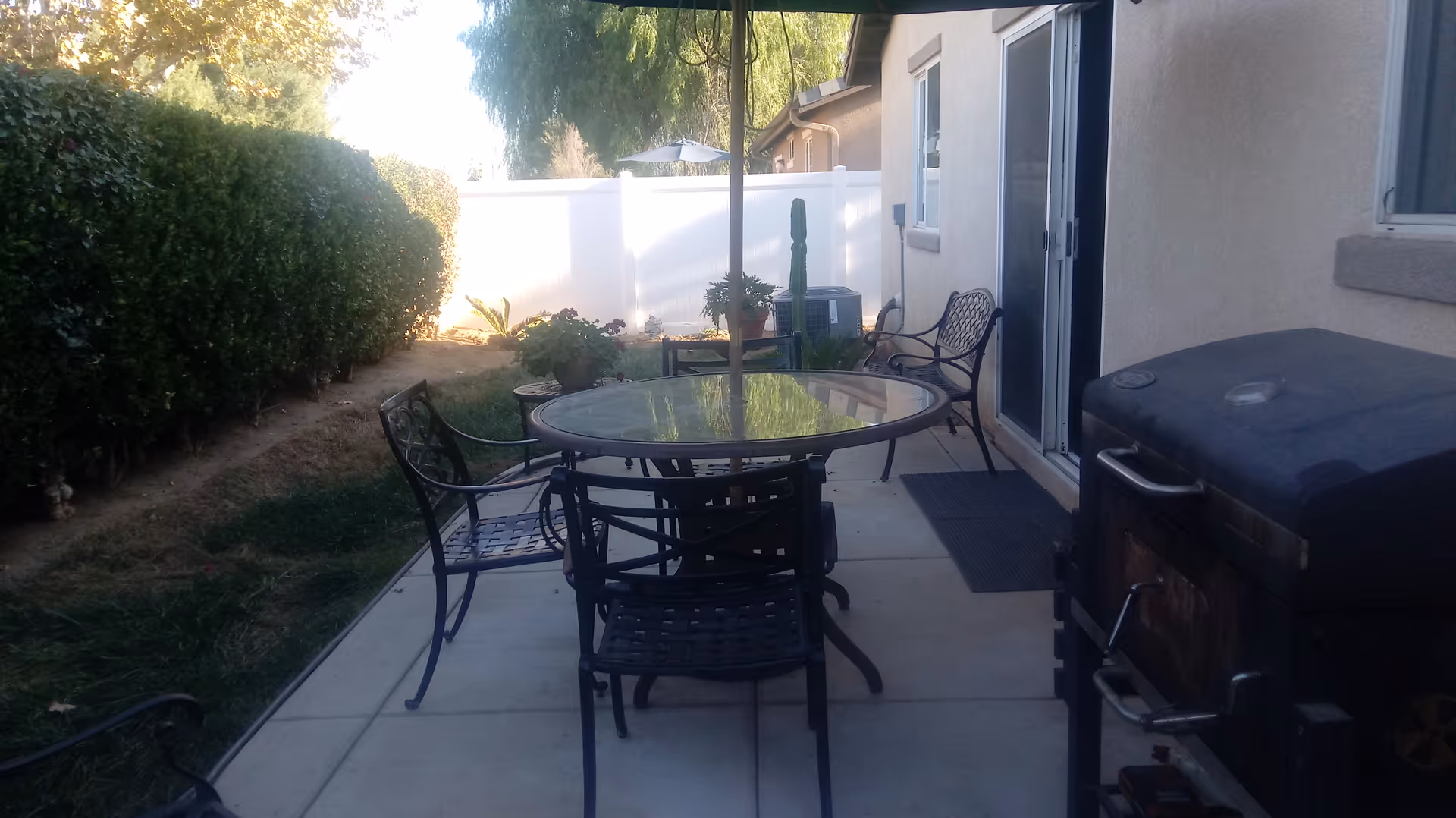 Backyard patio with a glass-top round table and metal chairs, a grill, sliding glass door to the house, hedge, and white fence.