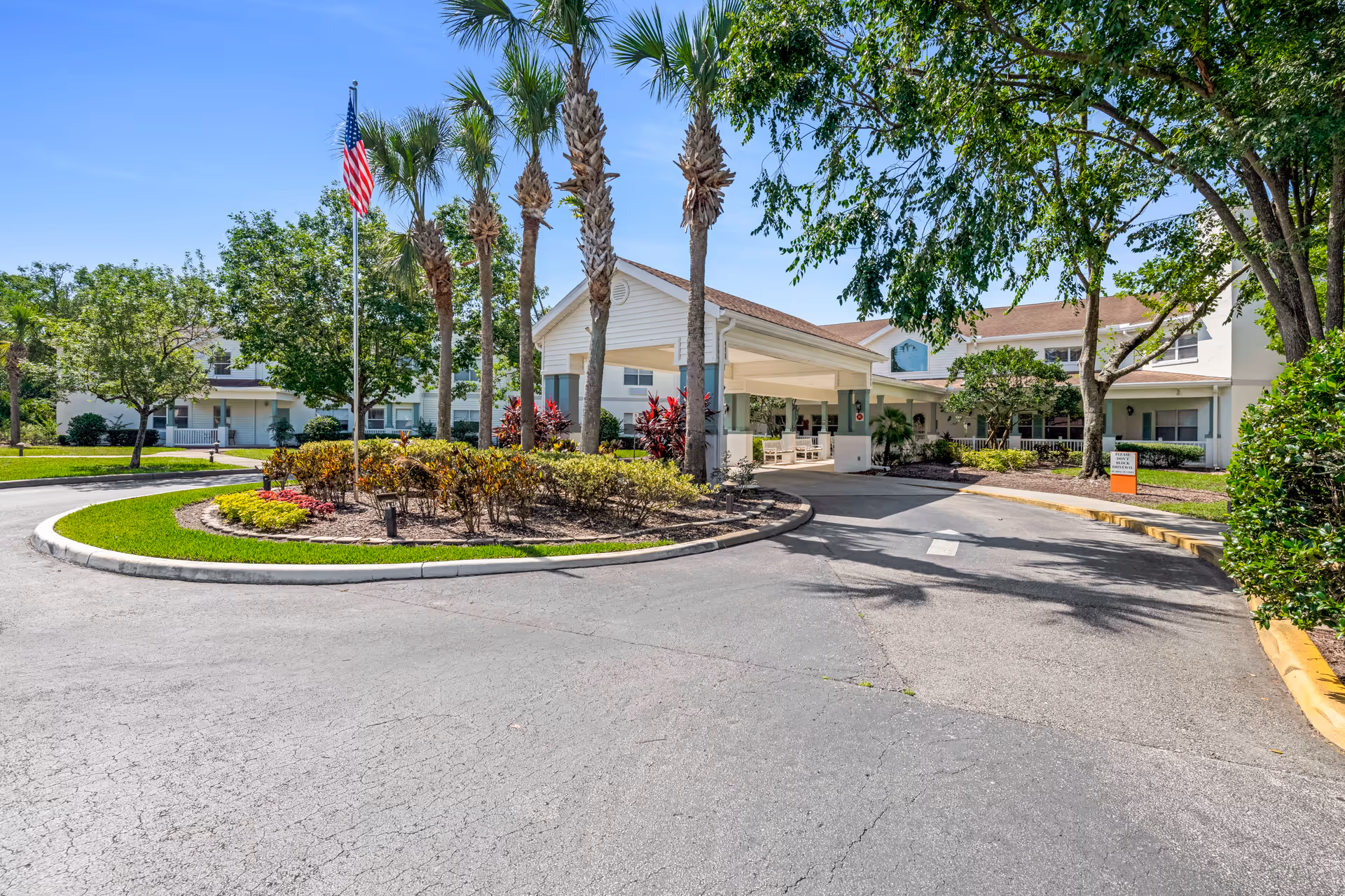 Exterior view of Ansley Parke at Orange City senior living facility entrance with a covered drop-off area, palm trees, an American flag, and well-maintained landscaping under a clear blue sky.