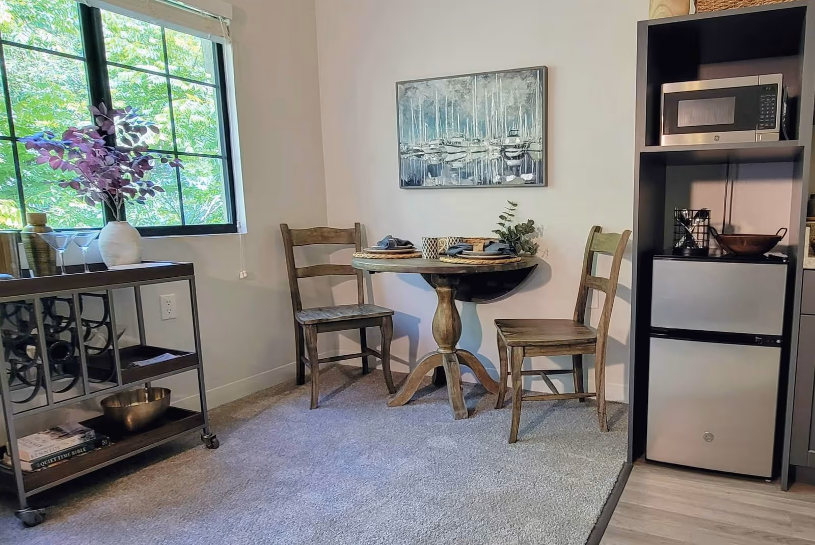 A small dining area with a round wooden table set for two with plates, napkins, and cups. Two wooden chairs are placed around the table. To the right, there is a shelving unit with a microwave on top and a mini refrigerator below. On the left, a window with a view of green trees lets in natural light. A metal cart with wine storage, a vase with purple flowers, and some books is positioned near the window. A painting of boats on water hangs on the wall above the table.