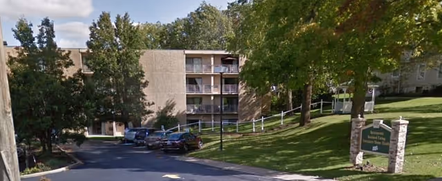 Exterior view of a multi-story senior living facility with balconies, surrounded by trees and a grassy area. Several cars are parked along the driveway leading to the building. A sign near the entrance reads 'St Martin Management'.
