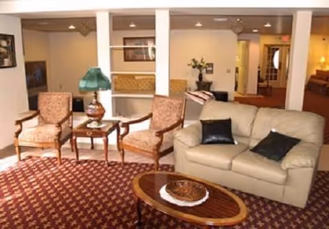 A cozy living room area with a beige leather sofa adorned with two black pillows, two patterned armchairs, a wooden coffee table with a decorative bowl, and a side table with a green lamp. The room has a patterned carpet and white pillars separating the space from an adjacent area with additional seating and decor.