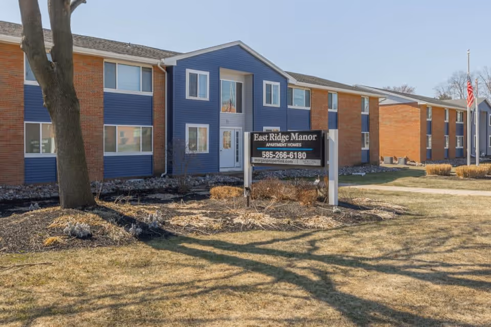 Exterior view of a two-story residential building with blue and brick siding. There is a large tree in the foreground and a sign in front that reads 'East Ridge Manor Apartment Homes' with a phone number and website. The lawn is dry with some landscaping around the sign, and an American flag is visible on the right side.