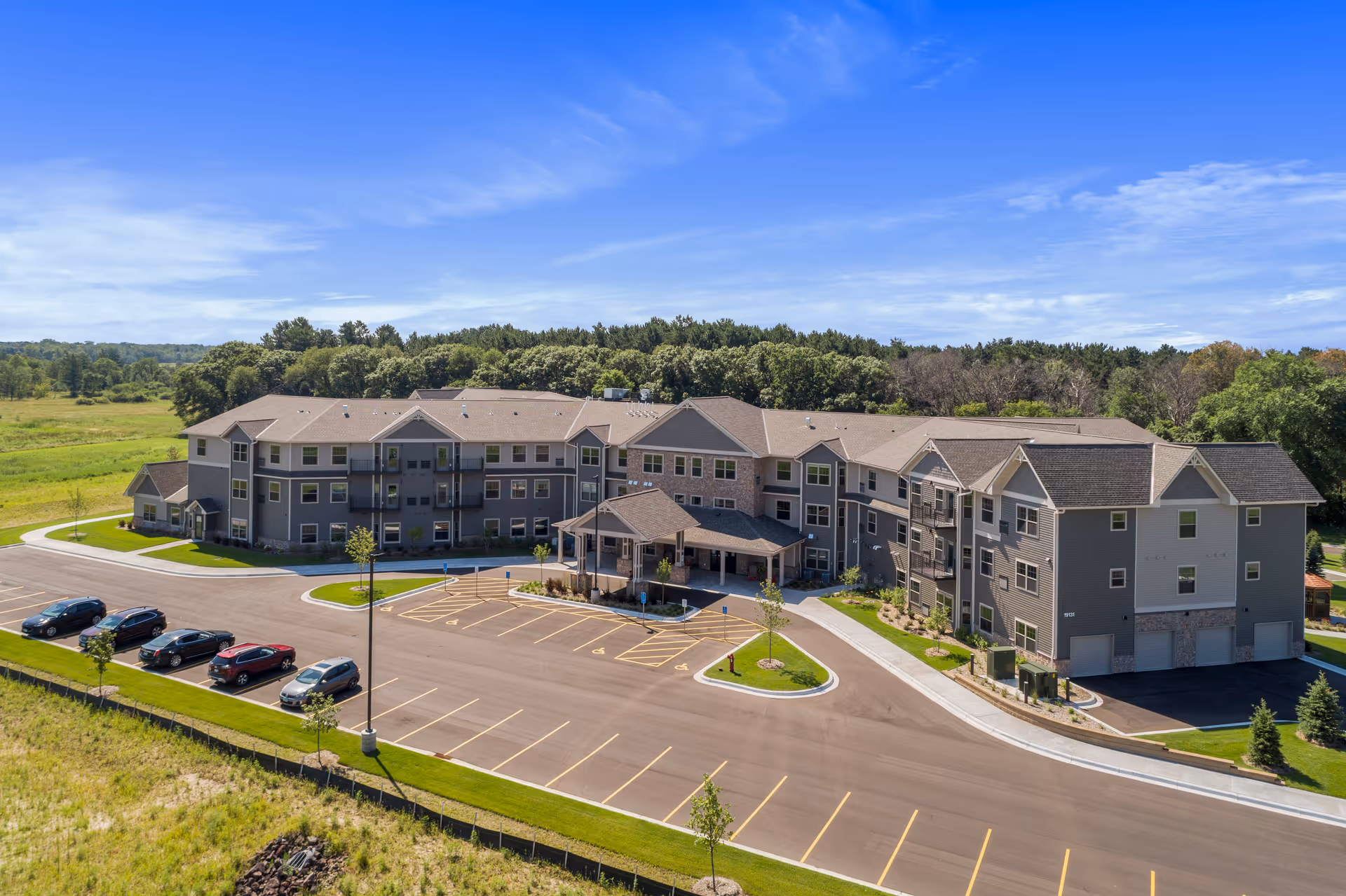 A large, modern three-story senior living facility building with a covered entrance and multiple balconies, surrounded by a parking lot with several cars and green landscaped areas, under a clear blue sky.