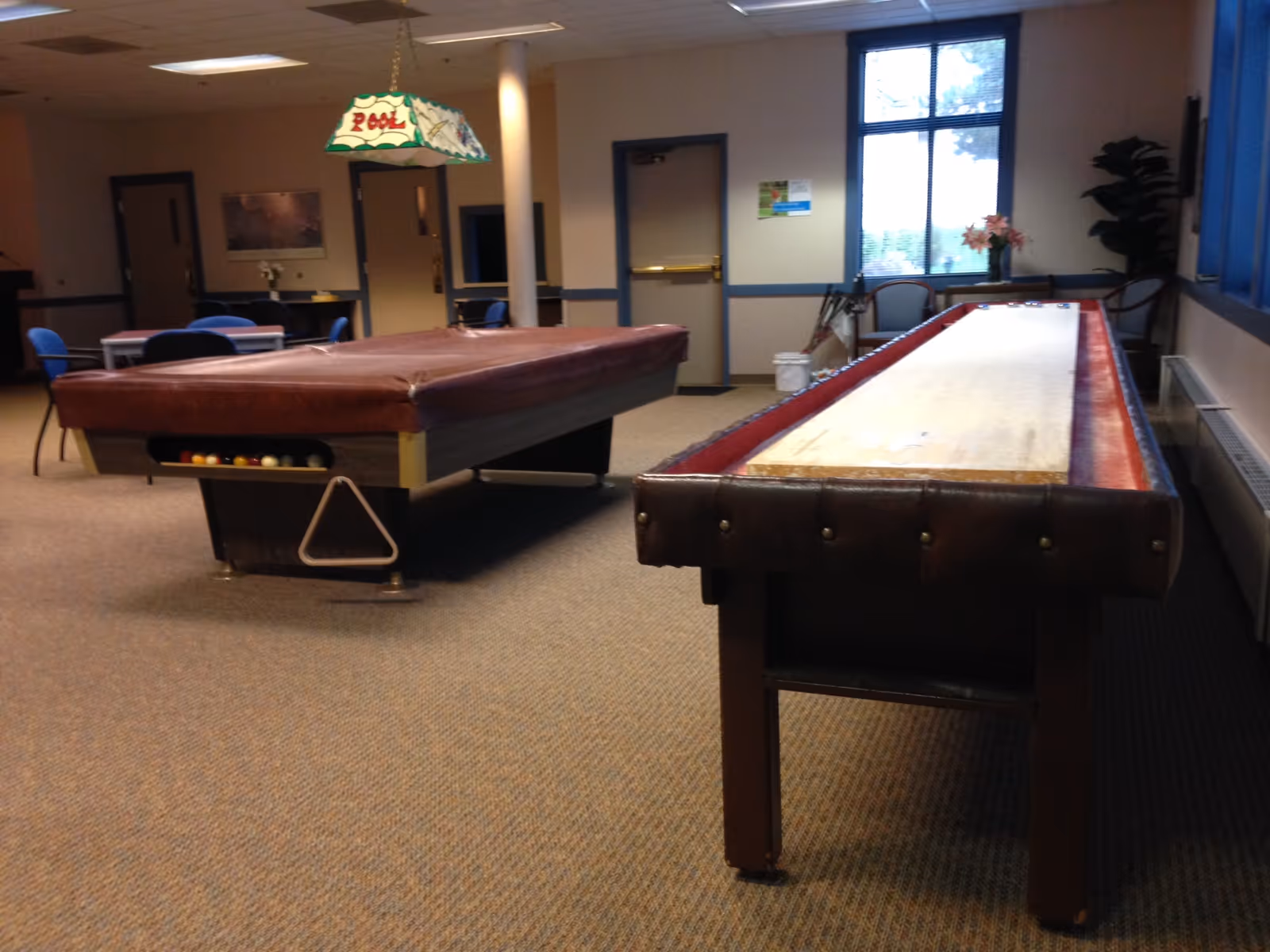 A recreational room interior with a covered pool table and a long shuffleboard table, chairs, and a hanging 'Pool' lamp.