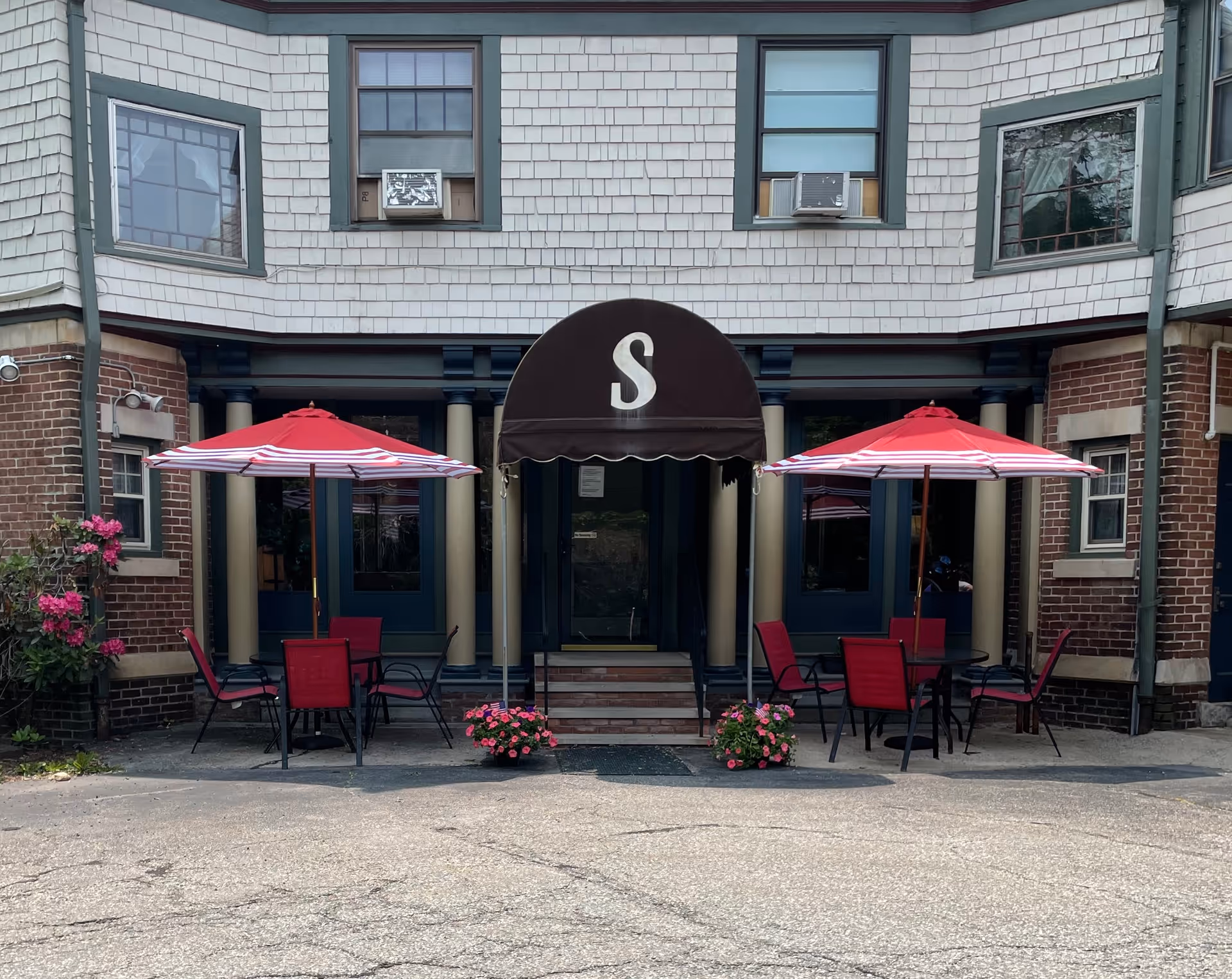 Front entrance of a building with a black awning displaying a large white letter 'S'. There are two red and white striped umbrellas shading outdoor tables with red chairs on either side of the entrance. Pink flowers in pots are placed near the steps leading to the door. The building has brick and shingle siding with several windows visible.