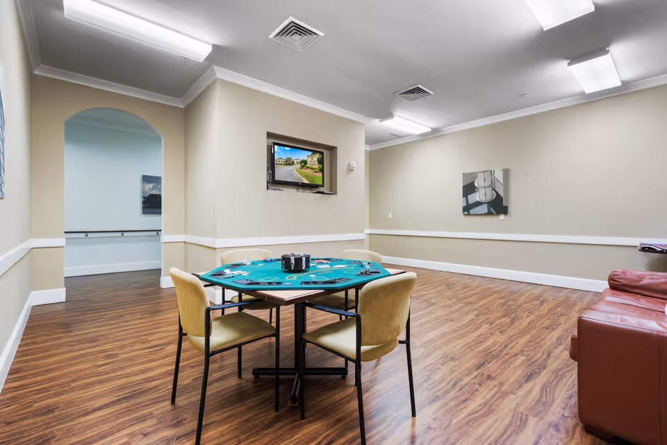 A spacious room with wood flooring featuring a green poker table with four chairs around it. A red leather couch is positioned against the right wall. The walls are beige with white trim, and there is a small TV mounted in a recessed wall niche displaying an outdoor scene. The room is well-lit with ceiling lights, and there is an arched doorway leading to another area.