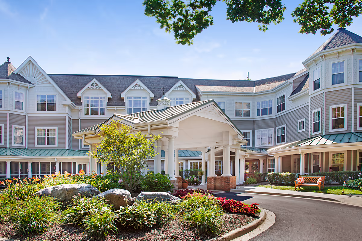 Exterior view of a senior living facility with a covered entrance, landscaped garden with rocks and plants, and a multi-story building with many windows under a clear blue sky.