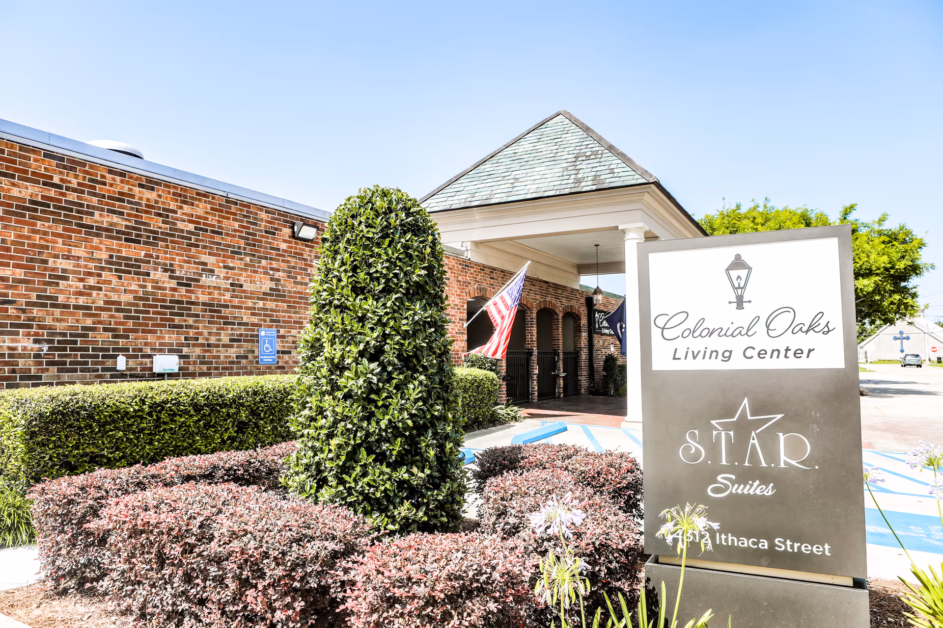 Entrance of Colonial Oaks Living Center showing the front sign, trimmed shrubs, and an American flag.