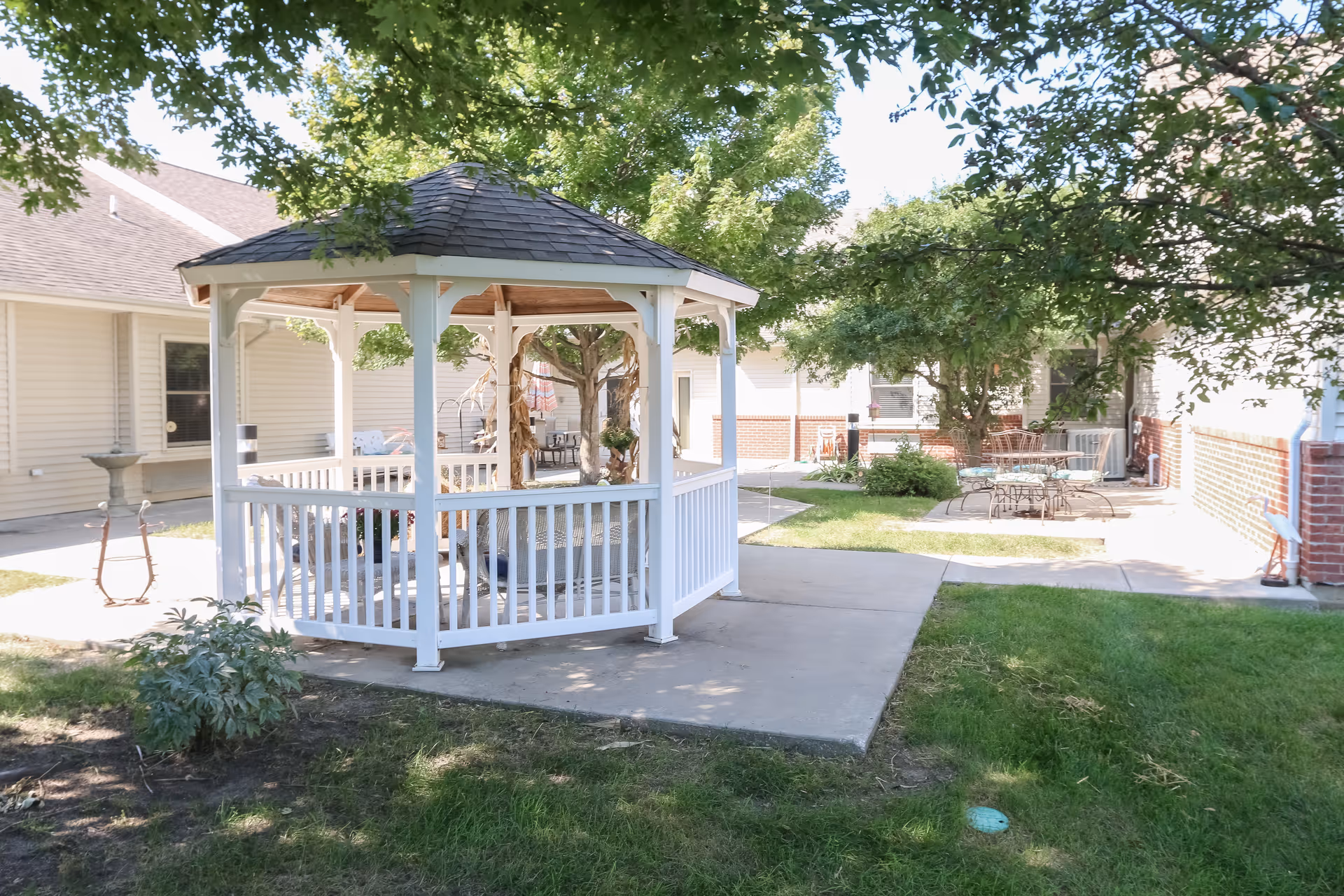 Outdoor courtyard area at Vintage Park at Paola featuring a white wooden gazebo with a shingled roof, surrounded by green grass, trees, and paved walkways. There are metal tables and chairs in the background near the building walls.