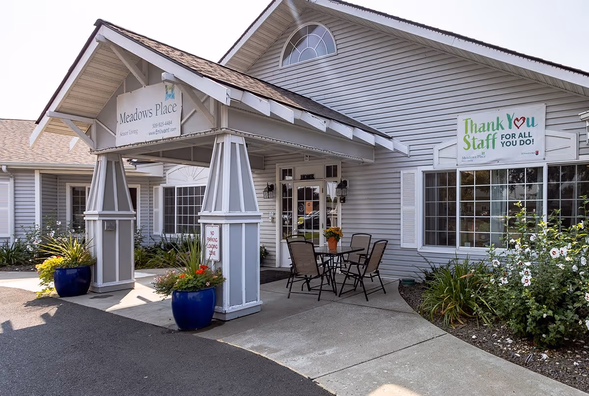 Exterior view of a senior living facility entrance with a covered porch supported by two large pillars. There are two large blue planters with flowers on either side of the entrance. A small outdoor seating area with a table and four chairs is visible near the door. The building is light gray with white trim and has a sign that reads 'Meadows Place Senior Living' above the entrance. Another sign on the building says 'Thank You Staff FOR ALL YOU DO!'.