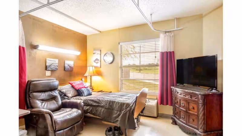 A nursing facility bedroom with a single bed, leather recliner, TV on a wooden dresser, window with blinds and pink curtains, and a bedside lamp.